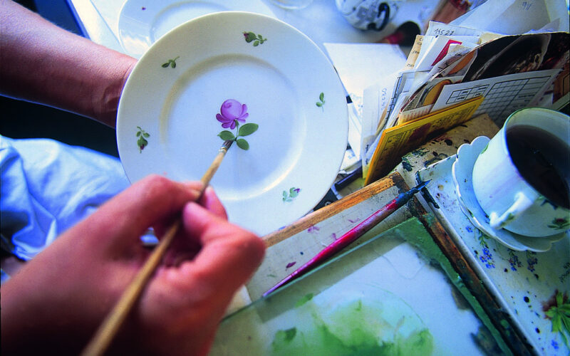 Eine Person malt mit einem dünnen Pinsel eine violette Blume mit grünen Blättern auf einen weißen Teller, in der Nähe befinden sich Malutensilien und eine Tasse Tee.