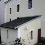 Ein blassgelbes Gebäude mit vergitterten Fenstern, einem kleinen Anbau, einem metallenen Räderwerk und an die Wand gelehnten Reinigungsgeräten.