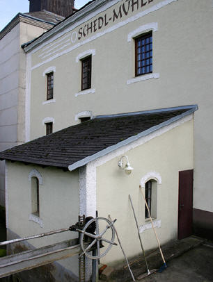 Ein blassgelbes Gebäude mit vergitterten Fenstern, einem kleinen Anbau, einem metallenen Räderwerk und an die Wand gelehnten Reinigungsgeräten.
