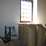 Große braune Papiersäcke sind an einer Wand neben einer altmodischen Waage unter einem Fenster mit farbigen Glasscheiben aufgereiht.