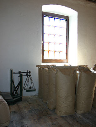 Große braune Papiersäcke sind an einer Wand neben einer altmodischen Waage unter einem Fenster mit farbigen Glasscheiben aufgereiht.