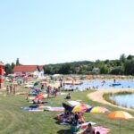 An einem sonnigen Tag entspannen sich Menschen auf Handtüchern und unter Sonnenschirmen an einem See. Im Hintergrund sind ein rot-weiß gestreifter Turm und mehrere kleine Gebäude zu sehen.