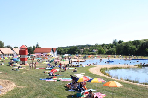An einem sonnigen Tag entspannen sich Menschen auf Handtüchern und unter Sonnenschirmen an einem See. Im Hintergrund sind ein rot-weiß gestreifter Turm und mehrere kleine Gebäude zu sehen.