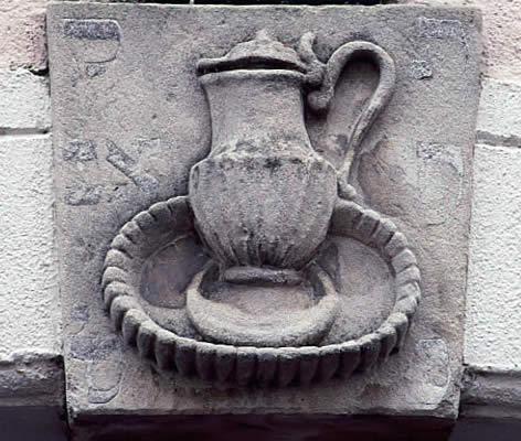 Steinrelief, das einen Krug und eine Schale auf einer rechteckigen Platte zeigt, umgeben von schwachen hebräischen Buchstaben, die in eine Ziegelmauer eingelassen sind.