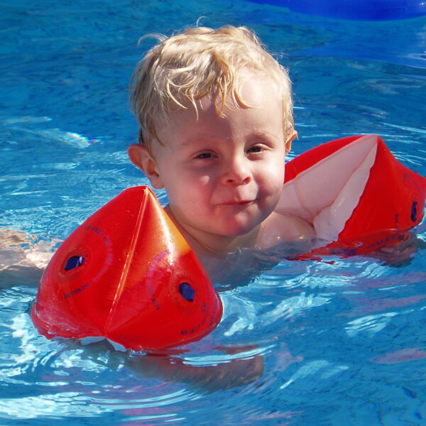 Junges Kind mit blondem Haar, das rote aufblasbare Armbänder trägt, schwimmt in einem Pool mit klarem blauen Wasser.