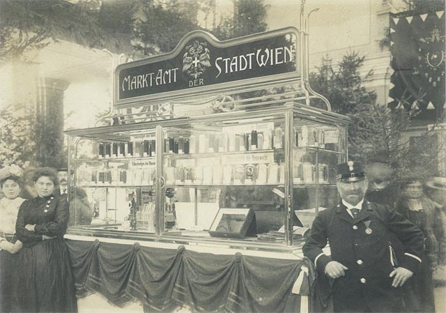 Ein alter Marktstand mit der Aufschrift "Marktamt der Stadt Wien", an dem Flaschen und Gläser ausgestellt sind, umgeben von Menschen, darunter ein Uniformierter im Vordergrund.