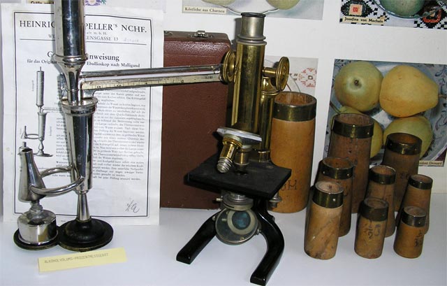 Ein altes Messingmikroskop wird zusammen mit wissenschaftlichen Instrumenten, beschrifteten Behältern und Papieren vor einem Hintergrund aus Postern und Äpfeln ausgestellt.
