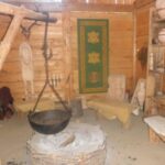 Ein rustikaler Holzraum mit einer zentralen Feuerstelle, einem Hängetopf, Bänken, Regalen mit Töpferwaren und einem grünen Wandteppich an der Wand.