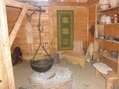 Ein rustikaler Holzraum mit einer zentralen Feuerstelle, einem hängenden Topf, Holzbänken, Regalen mit Töpferwaren und einem grünen Wandteppich an der Wand.