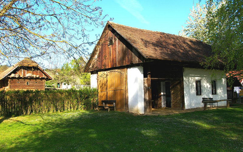 Ein rustikales Holzhaus mit schrägem Dach steht in einem grasbewachsenen Hof mit Bäumen und Sonnenlicht, neben einer Hecke und einer weiteren Holzkonstruktion im Hintergrund.