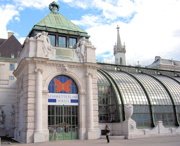 Gebäude aus Glas und Stein mit einem Schild mit der Aufschrift "Schmetterling Haus" über dem Eingang, mit einem Schmetterlingslogo und verschnörkelten architektonischen Details. Eine Person geht in der Nähe.