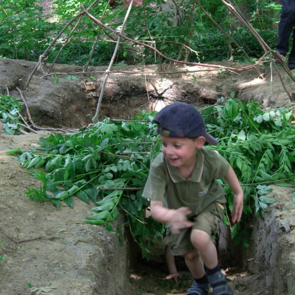 Ein kleiner Junge in grünem Outfit und Mütze rennt durch einen Erdgraben mit belaubten Zweigen und einer Rahmenkonstruktion aus Stöcken in einem Waldstück.