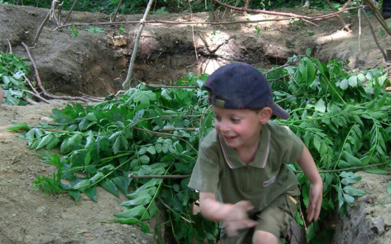 Ein kleiner Junge in grünem Outfit und Mütze rennt durch einen Erdgraben mit belaubten Zweigen und einer Rahmenkonstruktion aus Stöcken in einem Waldstück.
