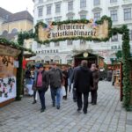 Menschen gehen durch den Eingang zum Altwiener Christkindlmarkt, einem Weihnachtsmarkt mit geschmückten Holzbuden auf einem Stadtplatz.