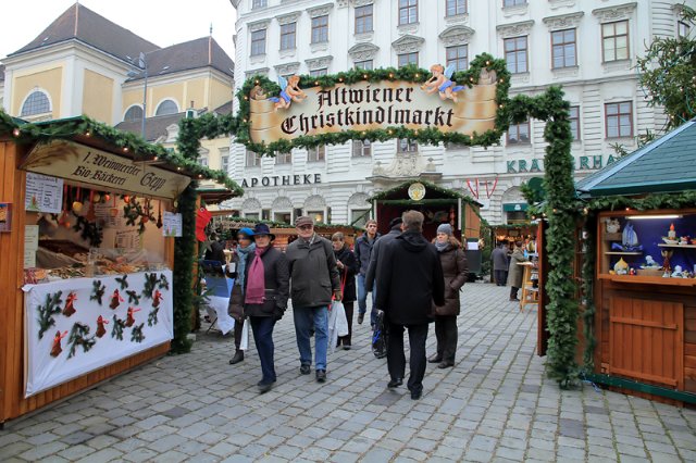 Menschen gehen durch den Eingang zum Altwiener Christkindlmarkt, einem Weihnachtsmarkt mit geschmückten Holzbuden auf einem Stadtplatz.
