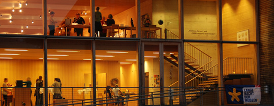 Blick durch große Glasfenster auf ein modernes Gebäude, in dem Menschen auf zwei Etagen interagieren, mit einem Treppenhaus und warmer Beleuchtung im Inneren.