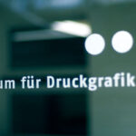 Glastür mit der Aufschrift "Museum für Druckgrafik" in deutscher Sprache, mit drei weißen Punkten über dem Wort "Museum" und einem unscharfen inneren Hintergrund.