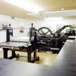 Eine Druckwerkstatt mit großen Druckmaschinen, einer Holzdecke, weißen Wänden, gerahmten Drucken und einer langen schwarzen Arbeitsplatte im Vordergrund.