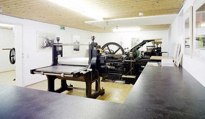 Eine Druckwerkstatt mit großen Druckmaschinen, einer Holzdecke, weißen Wänden, gerahmten Drucken und einer langen schwarzen Arbeitsplatte im Vordergrund.