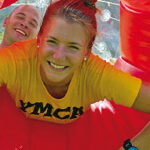 Zwei Personen lächeln, während sie sich in einem durchsichtigen, aufblasbaren Zorb-Ball befinden, wobei die vordere Person ein gelbes YMCA-Shirt trägt und rote Griffe hält.