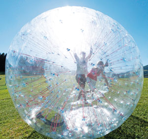 Zwei Personen in einem großen, durchsichtigen, aufblasbaren Ball, einem so genannten Zorb, rollen auf einer Wiese unter einem strahlend blauen Himmel.