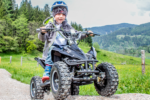 Ein kleines Kind mit Helm fährt mit einem kleinen vierrädrigen ATV auf einem unbefestigten Weg in einem grasbewachsenen, bergigen Außenbereich.
