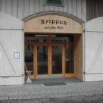 Eingang zu einem Gebäude mit großen Holztüren, Glaseingang und einem Schild darüber, auf dem „Krippen aus aller Welt“ steht. An der Fassade sind Hinweisschilder für WC und Museum zu sehen.