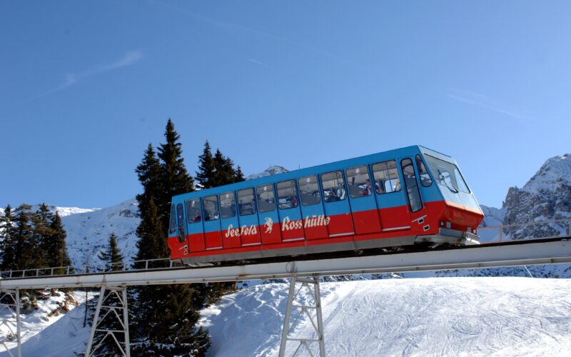 Eine rot-blaue Standseilbahn fährt auf einer erhöhten Strecke durch eine verschneite Berglandschaft unter einem klaren blauen Himmel.