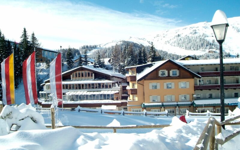 Schneebedeckte Gebäude und Kiefern stehen vor einem Berghang unter klarem Himmel; im Vordergrund sind mehrere senkrechte rote, gelbe und weiße Fahnen zu sehen.