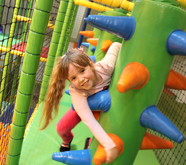 Ein junges Mädchen lächelt, während es auf ein großes, grünes, mit Stacheln versehenes Hindernis in einem von Netzen umgebenen Indoor-Spielbereich klettert.