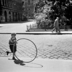 Ein kleines Kind rollt einen großen Reifen über eine Kopfsteinpflasterstraße, während im Hintergrund eine ältere Person auf dem Bürgersteig geht.