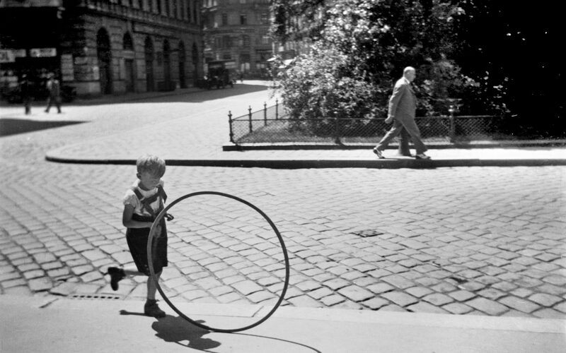 Ein kleines Kind rollt einen großen Reifen über eine Kopfsteinpflasterstraße, während im Hintergrund eine ältere Person auf dem Bürgersteig geht.