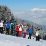 Eine Gruppe von Menschen in Winterkleidung steht mit erhobenen Skistöcken auf einem verschneiten Hang, umgeben von Bäumen und Bergen unter einem klaren Himmel.
