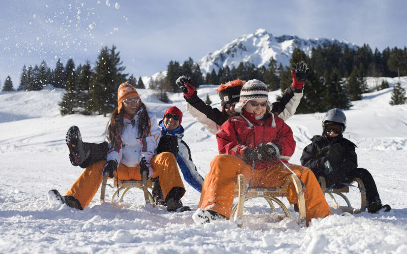 Sechs Personen in Winterkleidung fahren auf Holzschlitten einen verschneiten Hügel hinunter, im Hintergrund sind Bäume und ein schneebedeckter Berg zu sehen.