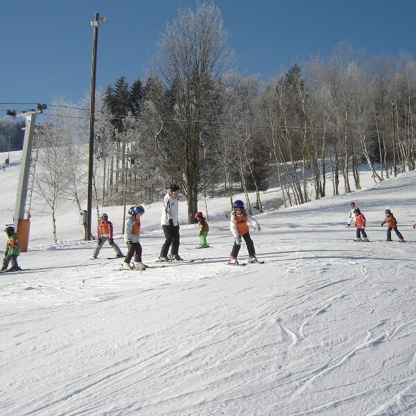 Eine Gruppe von Kindern in Skiausrüstung folgt einem Skilehrer an einem klaren Tag einen verschneiten Hang hinunter, mit Bäumen und einem Skilift im Hintergrund.