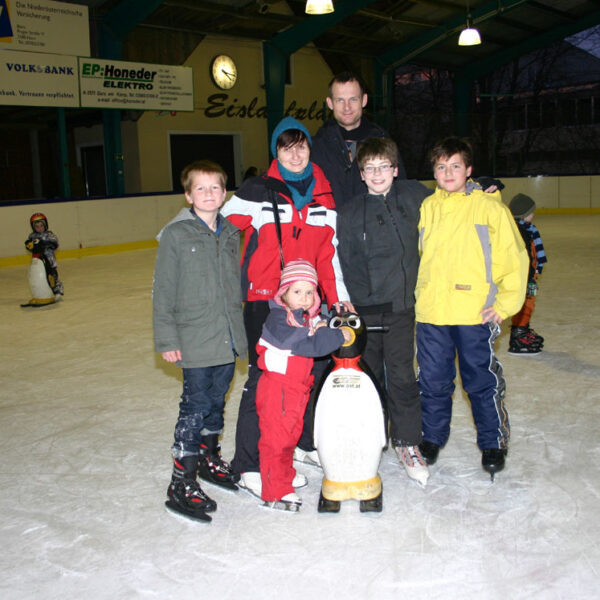 Sechs Kinder und ein Erwachsener posieren gemeinsam auf einer Eislaufbahn; ein Kind hält einen Pinguin als Eislaufhilfe.