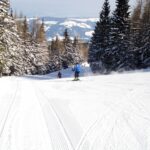 Zwei Personen fahren auf einer präparierten, schneebedeckten Piste, umgeben von schneebedeckten immergrünen Bäumen, unter einem klaren Himmel.