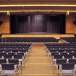 Leeres Auditorium mit blauen Stuhlreihen vor einer Bühne mit geschlossenen schwarzen Vorhängen, in einem modernen, gut beleuchteten Saal.