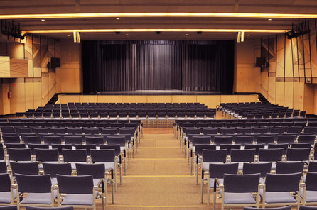 Leeres Auditorium mit blauen Stuhlreihen vor einer Bühne mit geschlossenen schwarzen Vorhängen, in einem modernen, gut beleuchteten Saal.