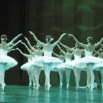 Balletttänzer in weißen Tutus treten auf der Bühne in einer koordinierten Formation auf, wobei ein männlicher Tänzer einer Gruppe von Tänzerinnen gegenübersteht.