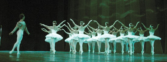 Balletttänzer in weißen Tutus treten auf der Bühne in einer koordinierten Formation auf, wobei ein männlicher Tänzer einer Gruppe von Tänzerinnen gegenübersteht.