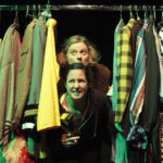 Zwei Personen blicken hinter einem Regal mit bunten und gemusterten Kleidungsstücken auf Kleiderbügeln hervor, beide mit ausdrucksstarker Miene, unter dramatischer Bühnenbeleuchtung.