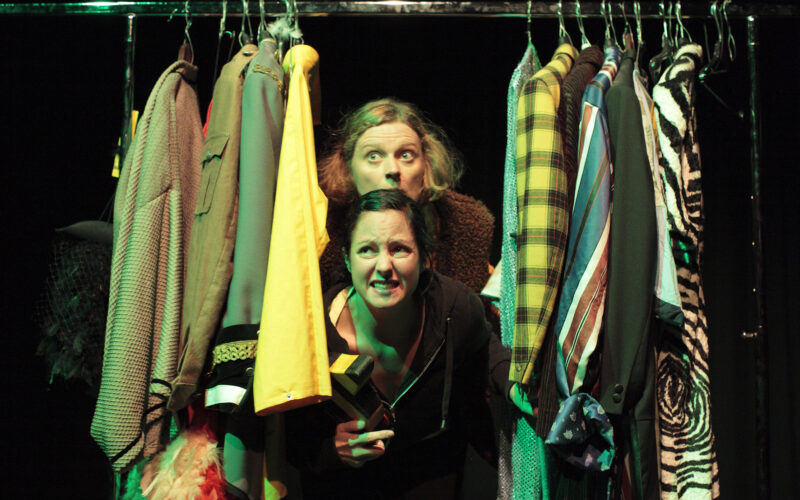 Zwei Personen blicken hinter einem Regal mit bunten und gemusterten Kleidungsstücken auf Kleiderbügeln hervor, beide mit ausdrucksstarker Miene, unter dramatischer Bühnenbeleuchtung.