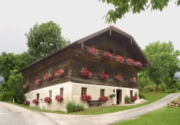 Ein zweistöckiges Haus mit hölzernen oberen Wänden und weißen unteren Wänden, verziert mit rosafarbenen Blumenkästen an den Fenstern, liegt zwischen zwei kurvenreichen Straßen, umgeben von Grün.