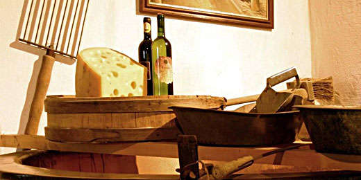 Ein großes Stück Käse, zwei Weinflaschen und rustikale Küchenutensilien sind auf einer hölzernen Fläche angeordnet, dahinter hängt ein gerahmtes Bild an der Wand.