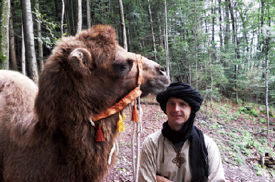 Ein Mann mit Kopftuch steht neben einem Kamel in einem Wald mit Bäumen und Grünpflanzen im Hintergrund.