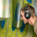 Ein Mann mit Kopfhörern lehnt sich an einen kleinen Bildschirm, der an einer bunten, gemusterten Wand angebracht ist.