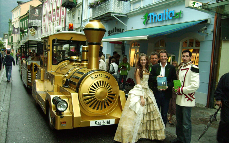 Eine Gruppe formell gekleideter Personen steht neben einem goldfarbenen Touristenzug in einer von Geschäften, darunter Thalia.at, gesäumten Straße in Bad Ischl, Österreich.