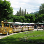 Ein kleiner gelber Touristenzug voller Fahrgäste fährt durch einen Park mit grünen Rasenflächen, Blumenbeeten und Bäumen. Im Hintergrund sind Marktstände und Menschen zu sehen.