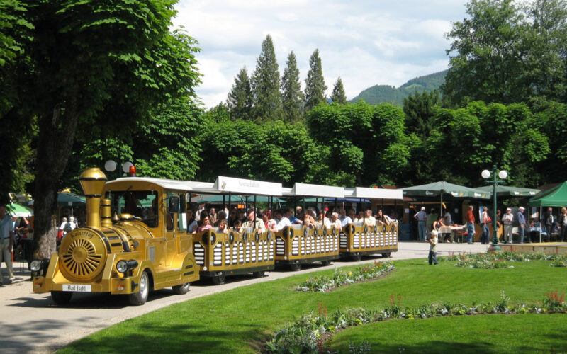 Ein kleiner gelber Touristenzug voller Fahrgäste fährt durch einen Park mit grünen Rasenflächen, Blumenbeeten und Bäumen. Im Hintergrund sind Marktstände und Menschen zu sehen.